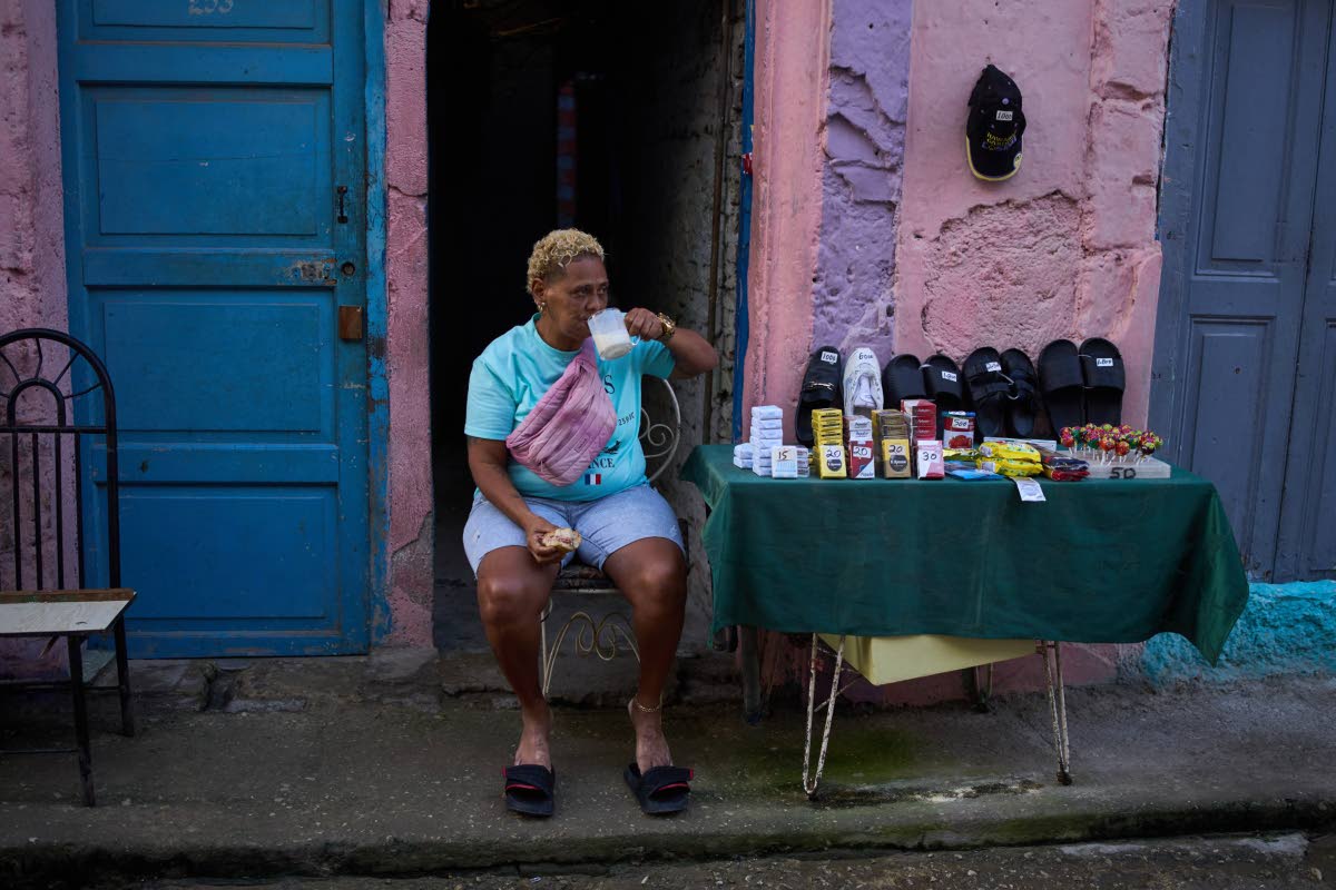 A vendor having breakfast sits by her table holding various products, from cigarettes to sandals, in Havana, Cuba.