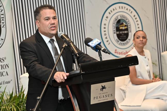 
Matthew Samuda, minister of water, environment, and climate change, speaks while Gabrielle Gilpin-Hudson, (right) president of Realtors Association of Jamaica, looks on at the Realtors Association of Jamaica Leaders in Real Estate Breakfast under the them